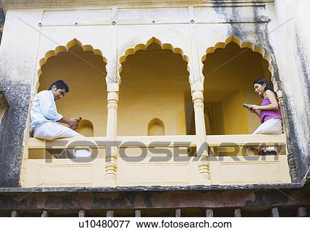 Stock Photo - Low angle view of a young couple sitting on the railing of a palace and operating mobile phones, Neemrana Fort Palace, Neemrana, Alwar, Rajasthan, India. Fotosearch