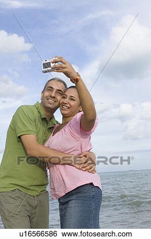 Stock Photograph - Mid adult couple taking a photograph of themselves on the beach. Fotosearch