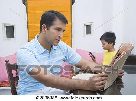 Stock Photography - Mid adult man reading a newspaper with his son holding a mobile phone in the background. Fotosearch
