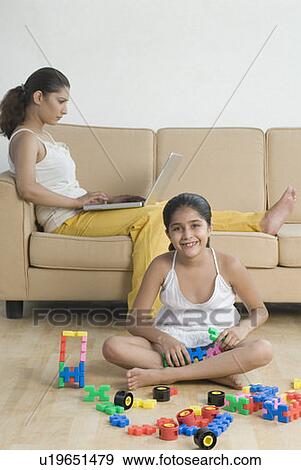 Stock Photo - Portrait of a girl playing with building blocks and her mother using a laptop behind her. Fotosearch