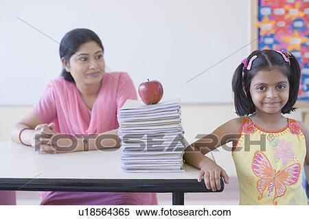Stock Photography - Portrait of a schoolgirl smirking with a teacher sitting at a desk behind her. Fotosearch