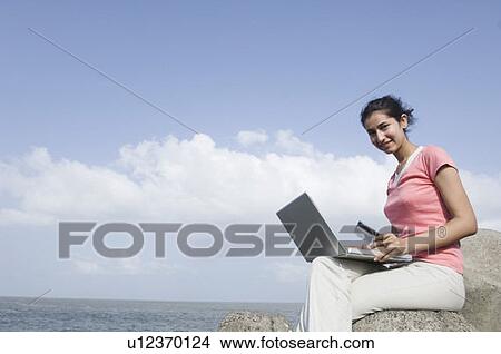 Picture - Portrait of a young woman sitting on a rock and working on a laptop . Fotosearch