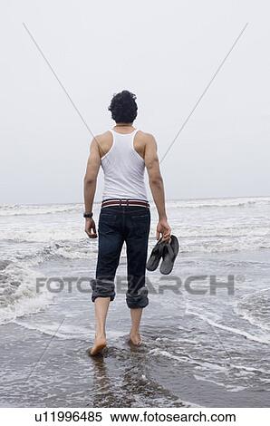 Stock Photography - Rear view of a young man walking on the beach with holding a pair of flip-flops. Fotosearch