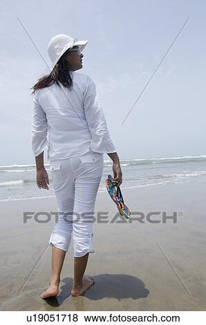 Rear view of a young woman holding flip-flop and walking on the beach View Large Photo Image Stock Photo - Rear view of a young woman holding flip-flop and walking on the beach. Fotosearch