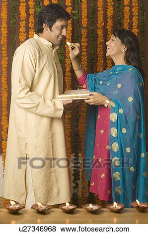 Stock Photo - Side profile of a young woman feeding a piece of burfi to a mid adult woman and smiling. Fotosearch