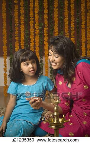 Stock Image - Side profile of a young woman holding a traditional diwali lamp and her daughter looking at her. Fotosearch