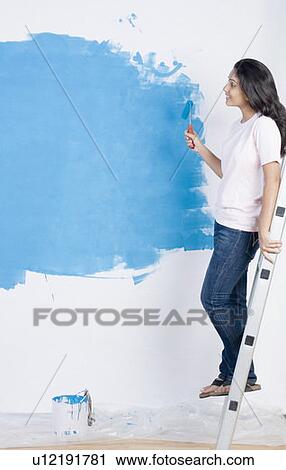 Stock Image - Side profile of a young woman painting a wall with a paint roller and leaning on a step ladder. Fotosearch