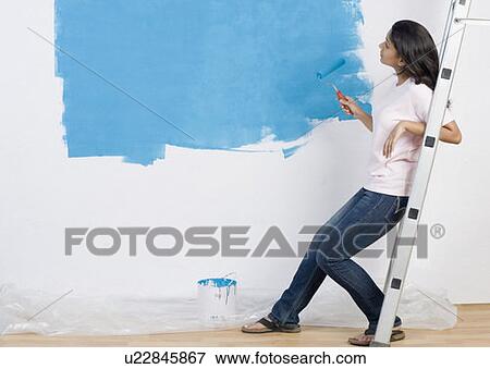 Stock Photo - Side profile of a young woman painting a wall with a paint roller and leaning on a step ladder. Fotosearch