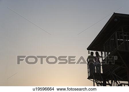 Picture - Silhouette of a man and a woman standing on the beach hut. Fotosearch