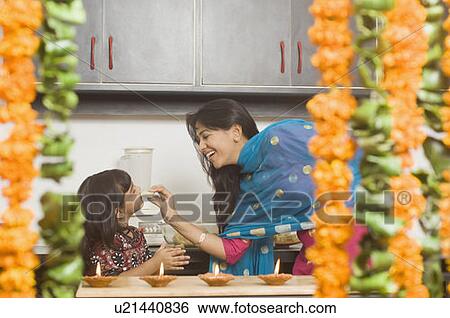 Young woman feeding a piece of burfi to her daughter in a domestic kitchen and smiling View Large Photo Image Stock Photograph - Young woman feeding a piece of burfi to her daughter in a domestic kitchen and smiling. Fotosearch