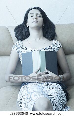 Stock Image - Young woman sitting on a couch with her eyes closed and holding a book. Fotosearch
