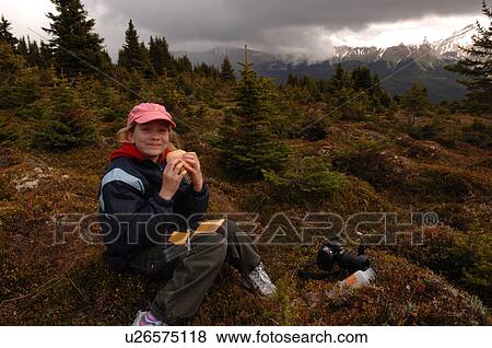 Stock Photo - Girl eating while sitting on ground in forest, Jasper National Park, Canada. Fotosearch
