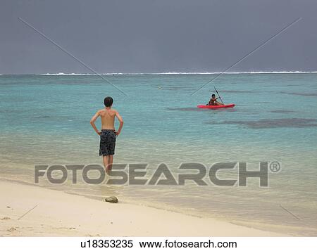 Stock Photography - Rear of a young man standing on a beach watching a boat on the sea. Fotosearch