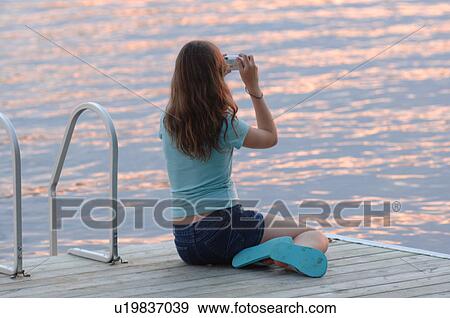 Teenage girl sitting on a dock taking a picture View Large Photo Image Stock Photo - Teenage girl sitting on a dock taking a picture. Fotosearch