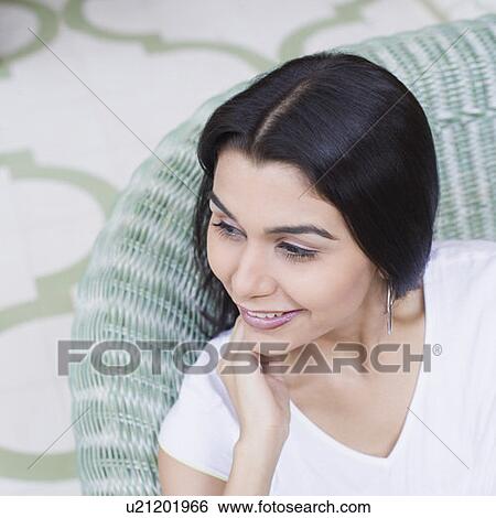 Close-up of a young woman sitting on a couch with her hand on her chin and smiling View Large Photo Image Stock Photograph - Close-up of a young woman sitting on a couch with her hand on her chin and smiling. Fotosearch