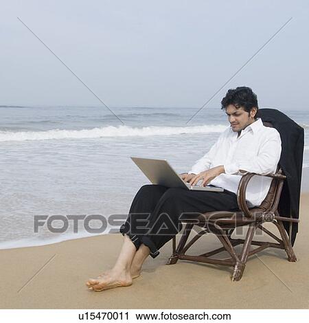 Stock Image - Businessman sitting in an armchair and using a laptop on the beach. Fotosearch