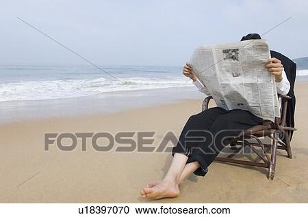 Stock Image - Businessman sitting in an armchair and reading a newspaper on the beach. Fotosearch