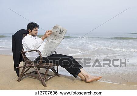 Stock Photograph - Businessman sitting in an armchair and reading a newspaper on the beach. Fotosearch