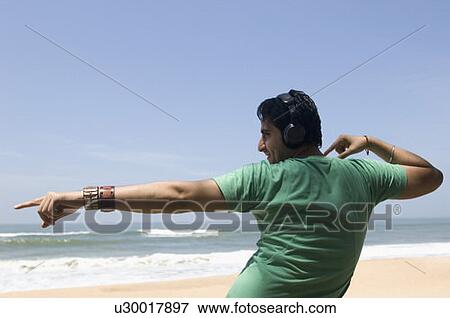 Stock Photo - Rear view of a young man wearing headphones and dancing on the beach. Fotosearch
