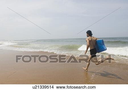 Side profile of a young man carrying a surfboard and running on the beach View Large Photo Image Picture - Side profile of a young man carrying a surfboard and running on the beach. Fotosearch