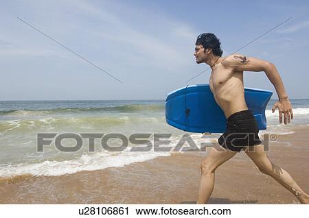 Stock Image - Young man carrying a surfboard and running on the beach. Fotosearch