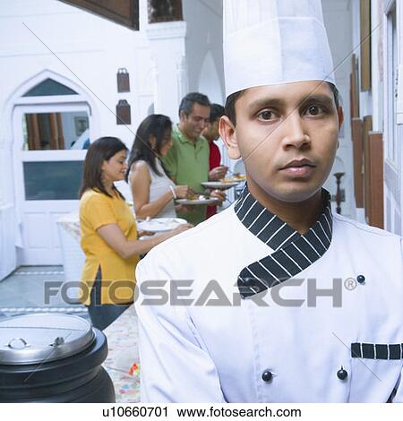 Stock Image - Portrait of a chef with two mid adult couples having lunch in the background. Fotosearch