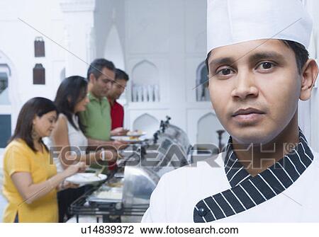 Stock Image - Portrait of a chef with two mid adult couples having lunch in the background. Fotosearch