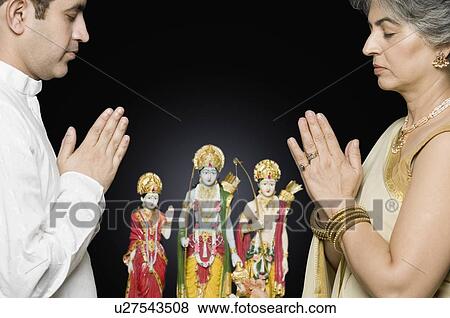 Stock Photo - Side profile of a mature woman and her son praying in front of figurines of God. Fotosearch