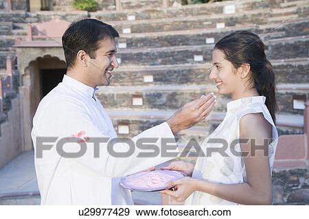 Stock Photo - Side profile of a mid adult man applying powder paint on the face of a young woman, Neemrana Fort Palace, Neemrana, Alwar, Rajasthan, India. Fotosearch