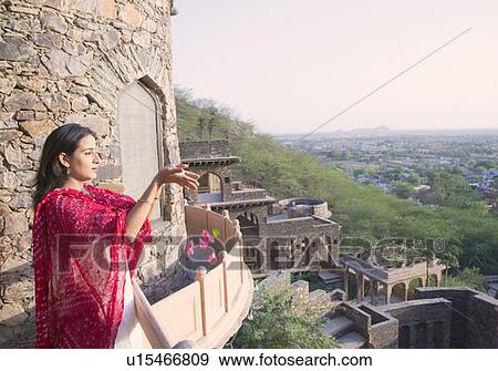 Stock Photo - Side profile of a mid adult woman standing at a balcony and offering flowers, Neemrana Fort Palace, Neemrana, Alwar, Rajasthan, India. Fotosearch