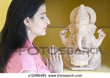 Stock Photography - Side profile of a mid adult woman praying in front of a statue of God Ganesha. Fotosearch