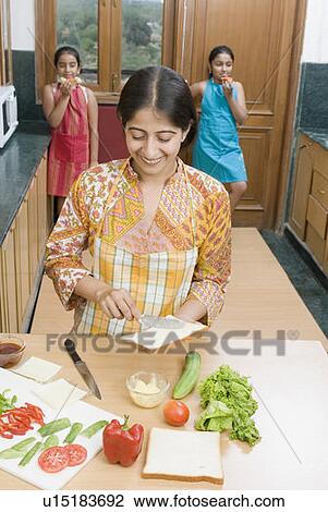 Stock Image - Mid adult woman spreading butter on a bread slice with her daughters in the background. Fotosearch