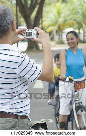 Rear view of a mid adult man taking a photograph of his son and wife riding a bicycle View Large Photo Image Stock Photo - Rear view of a mid adult man taking a photograph of his son and wife riding a bicycle . Fotosearch