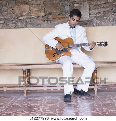 Young man sitting on a bench and playing a guitar View Large Photo Image Stock Photograph - Young man sitting on a bench and playing a guitar. Fotosearch