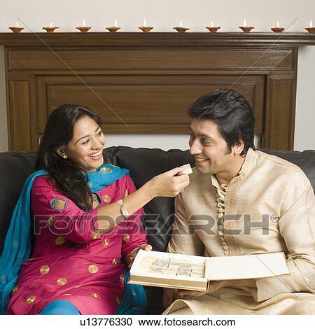 Stock Image - Close-up of a young woman feeding a piece of burfi to a mid adult man and smiling. Fotosearch