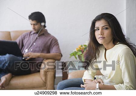 Stock Photo - Portrait of a young woman sitting in an armchair with a young man using a laptop in the background. Fotosearch