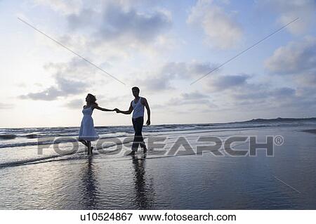 Stock Photo - Young couple holding each other's hands and walking on the beach. Fotosearch