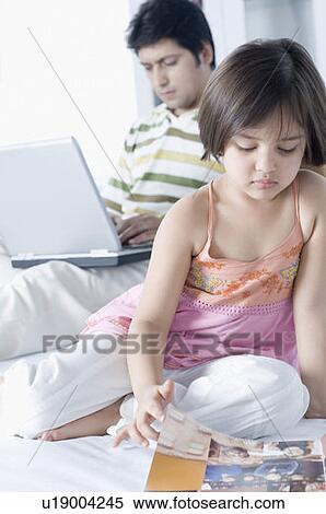 Stock Photography - Close-up of a girl reading a magazine with her father working on a laptop behind her. Fotosearch