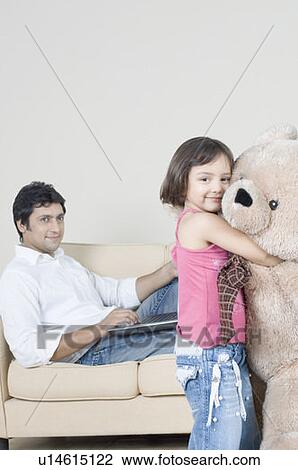 Stock Image - Portrait of a girl hugging a teddy bear with her father using a laptop beside her. Fotosearch