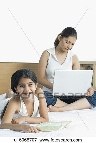 Stock Photo - Portrait of a girl lying on the bed and her mother using a laptop. Fotosearch