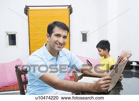 Stock Image - Portrait of a mid adult man holding a newspaper with his son sitting beside him. Fotosearch