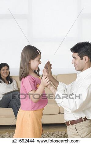 Picture - Side profile of a girl playing with her father and her mother sitting on a couch in the background. Fotosearch