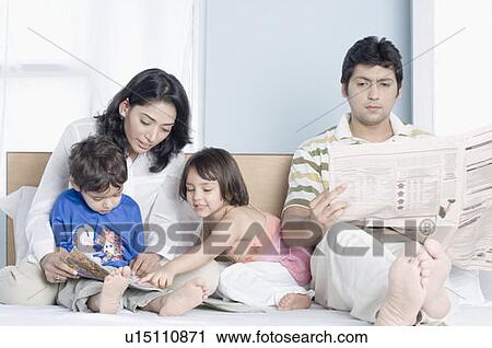 Stock Image - Young woman teaching her children with her husband sitting beside them and reading a newspaper. Fotosearch