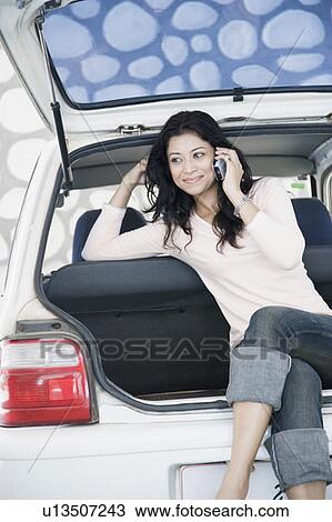 Stock Image - Young woman sitting in a car trunk and talking on a mobile phone. Fotosearch