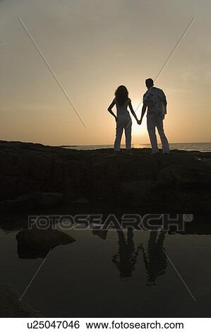 Rear view of a couple holding hands of each other and standing on the coast View Large Photo Image Stock Photograph - Rear view of a couple holding hands of each other and standing on the coast. Fotosearch