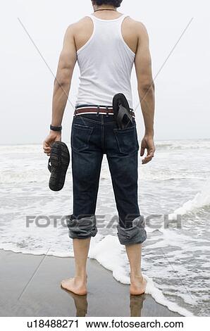 Rear view of a young man standing on the beach with holding a flip-flop View Large Photo Image Stock Image - Rear view of a young man standing on the beach with holding a flip-flop. Fotosearch