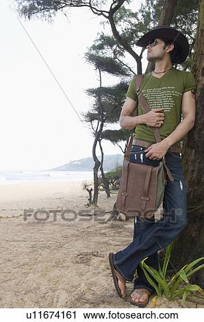 Stock Image - Young man carrying a shoulder bag and leaning against a tree trunk on the beach. Fotosearch