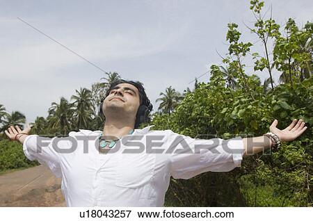 Young man standing with his arms outstretched and listening to music View Large Photo Image Stock Photo - Young man standing with his arms outstretched and listening to music. Fotosearch