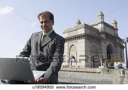 Stock Photography - Businessman using a laptop with a monument in the background, Gateway of India, Mumbai, Maharashtra, India. Fotosearch