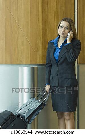 Picture - Portrait of a businesswoman talking on a mobile phone and holding her luggage. Fotosearch